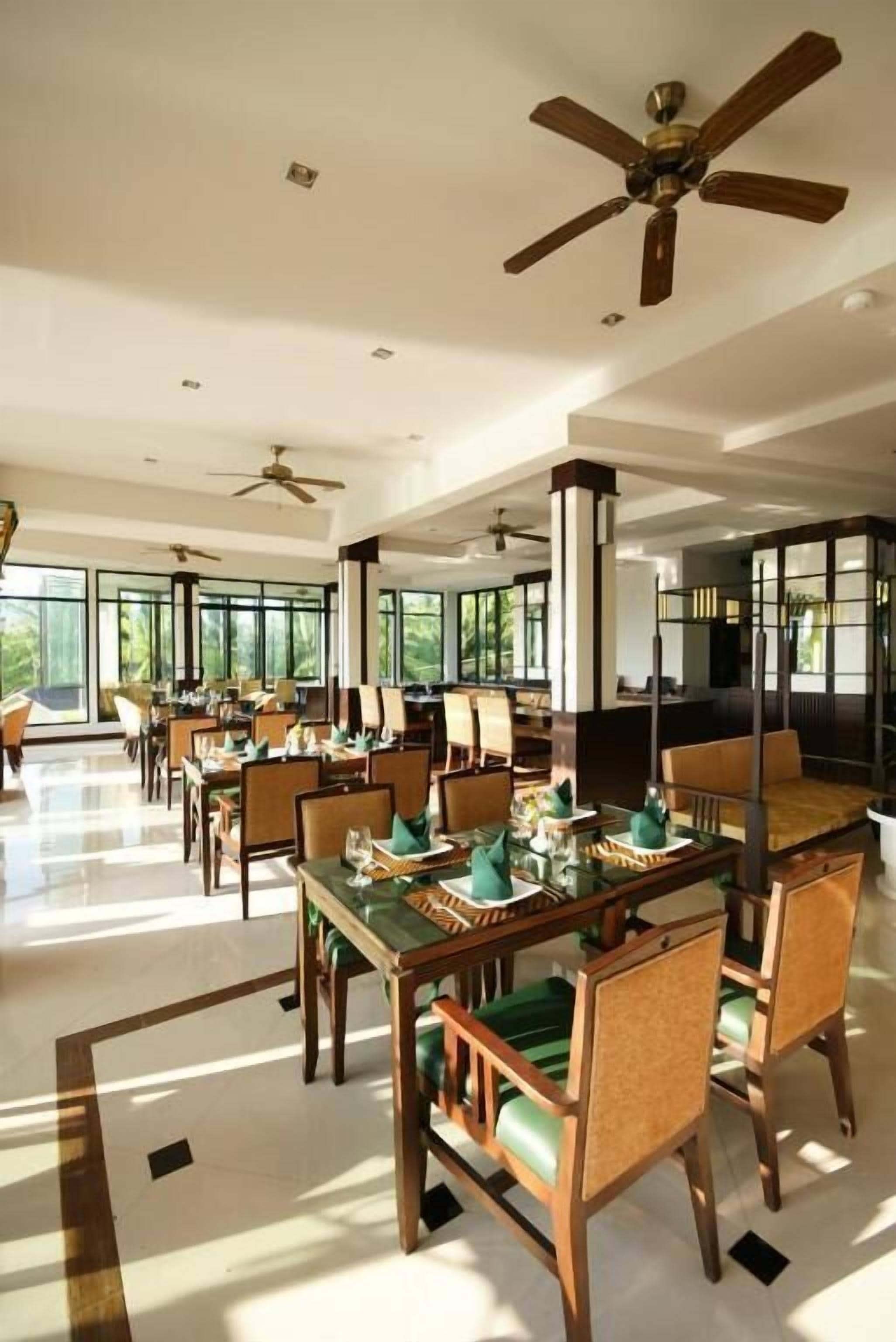 Un vaste espace dédié à la salle à manger, avec de nombreuses tables et chaises, des ventilateurs de plafond et de grandes fenêtres qui laissent entrer la lumière naturelle et offrent une vue agréable.