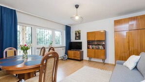 A living room with a dining table, chairs, a sofa, a TV, wooden cabinets, and a window with curtains. - ShortStayPoland Piekałkiewicza B112 (Warsaw)