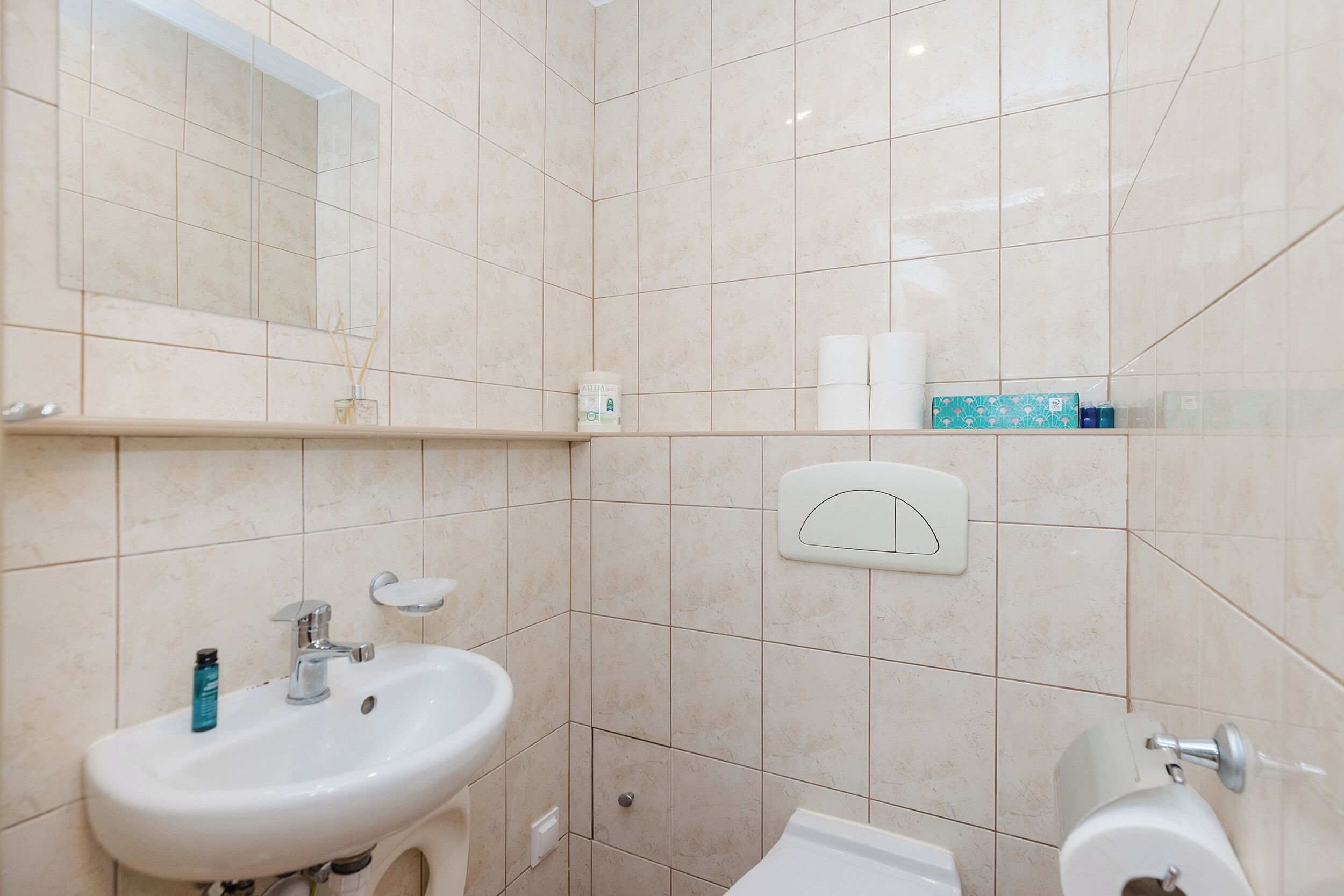 Un baño con lavamanos, espejo, inodoro y repisas con artículos de aseo.