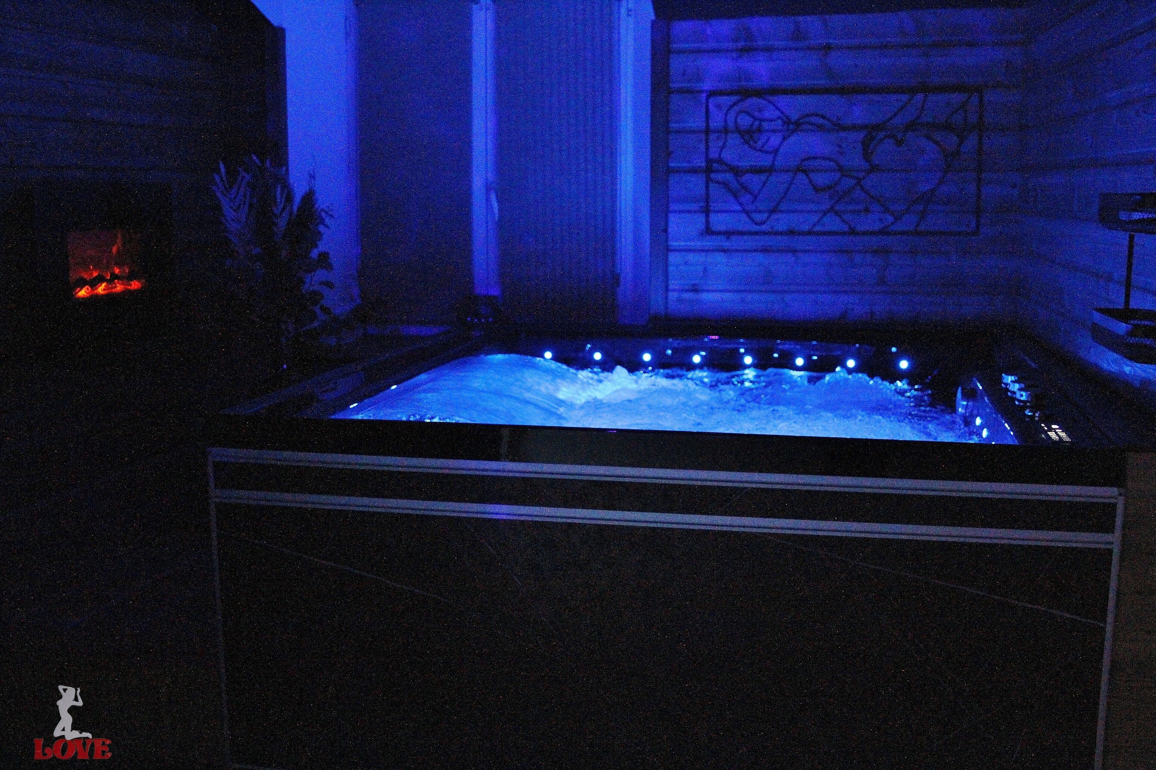 Een wellnessruimte met een bubbelbad verlicht met blauw licht, een open haard en een muur met een geometrisch ontwerp.