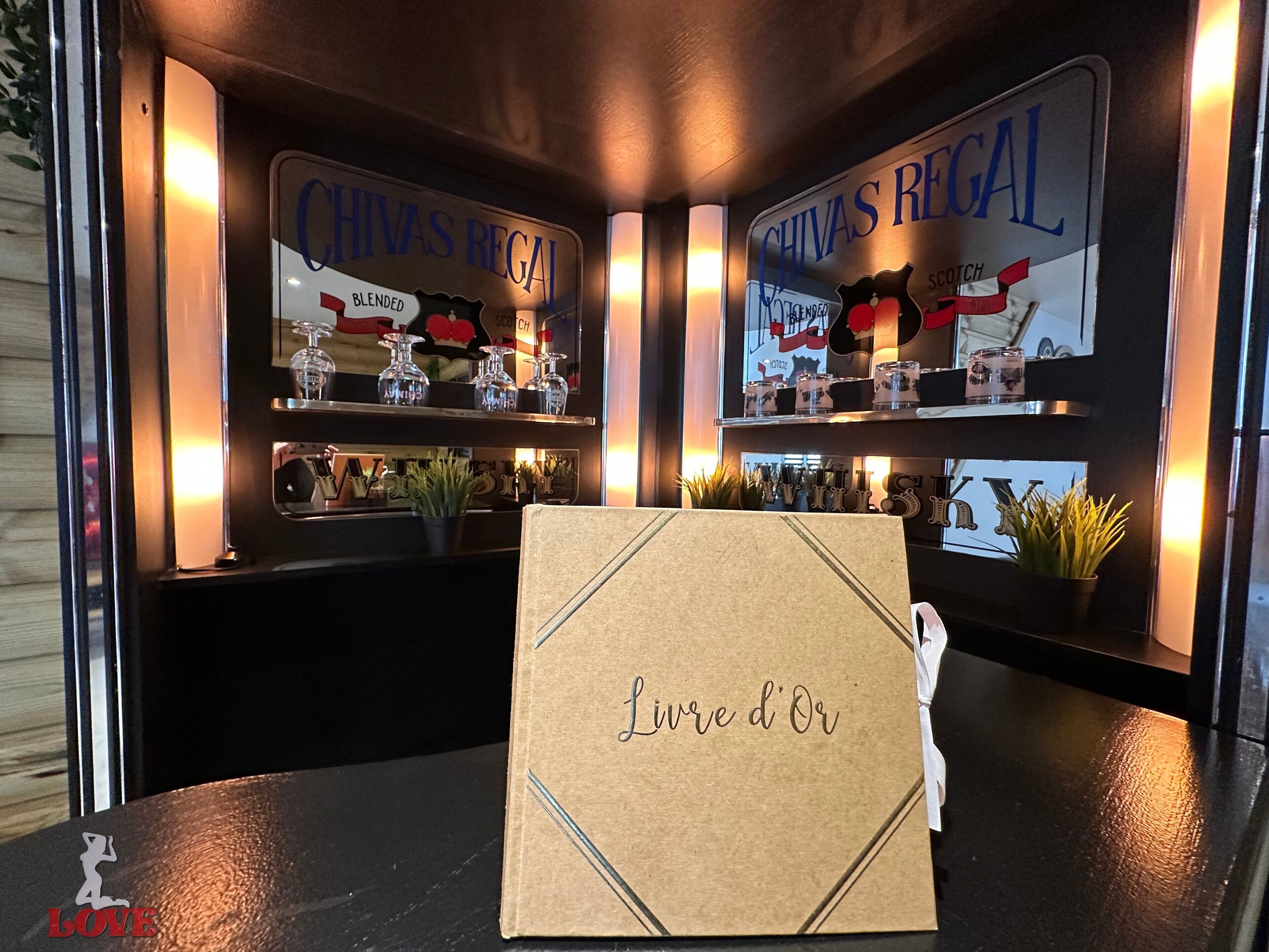 Un coin bar agrémenté d’enseignes « Chivas Regal », d’une enseigne « Blended Scotch » et d’une carte « Love d’Or » posée sur une table.