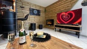 Een knus vertrek met een houten wand, een open haard, een televisie met een hartvormig scherm, een fles champagne en glazen op een houten tafel.
