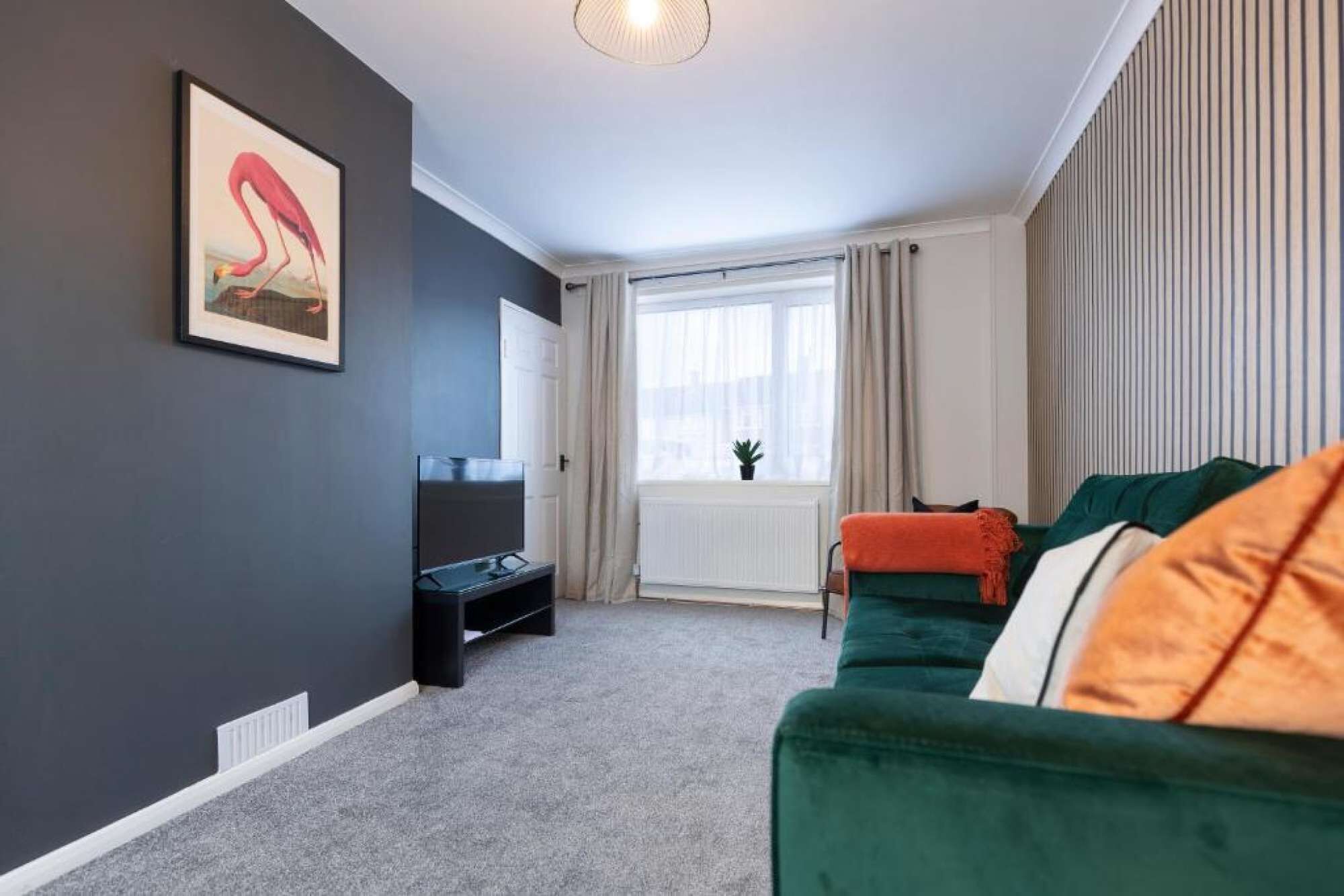 Ein Wohnzimmer mit dunkler Wand, einem gerahmten Bild, einem Flachbildfernseher, einem grünen Sofa mit orangenen Kissen und einem Fenster mit Vorhängen.