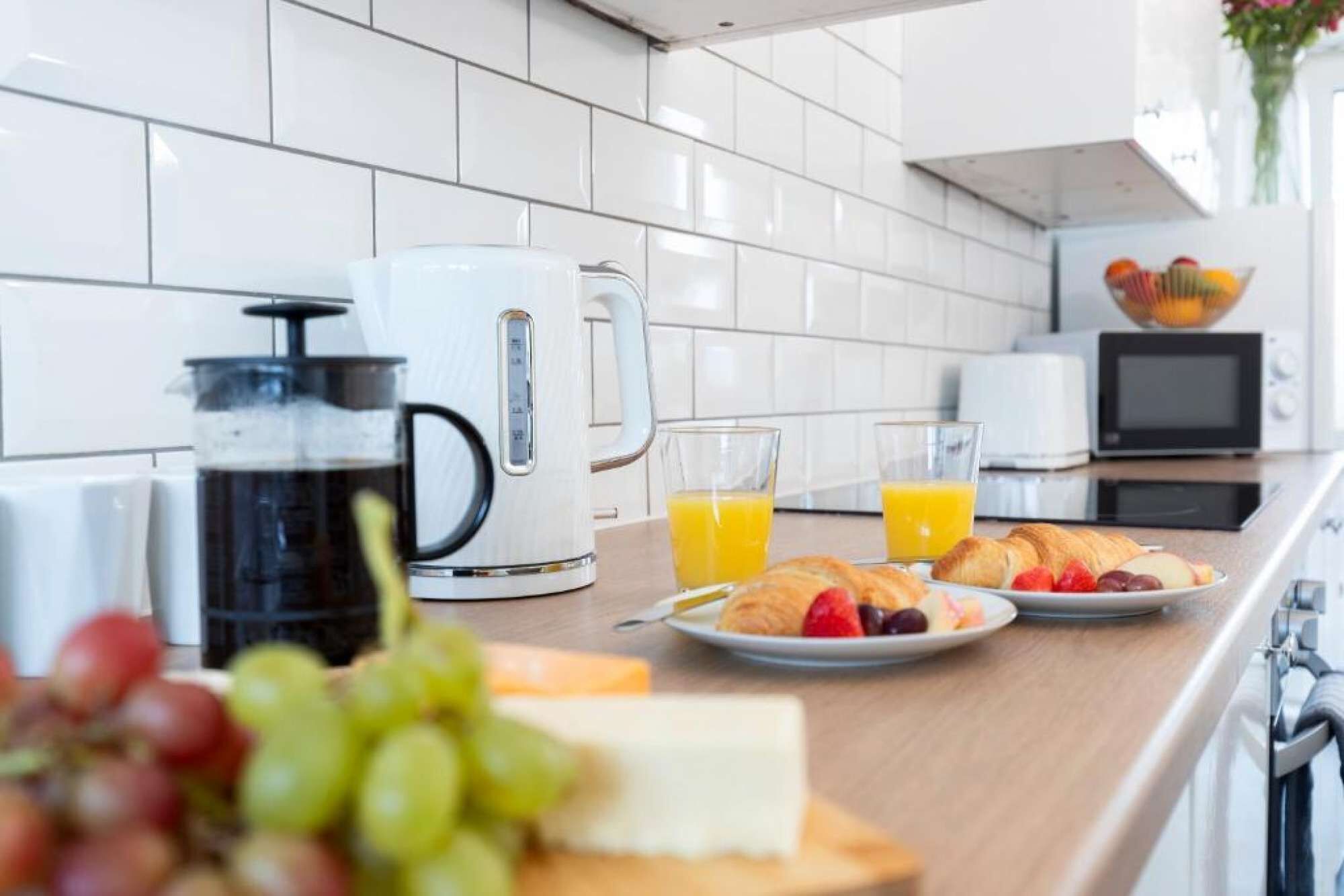 A modern kitchen with white tiles, a coffee maker, a kettle, a toaster, and a fruit bowl.