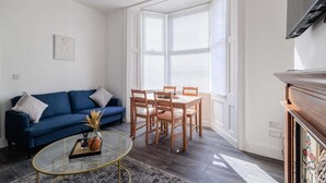 A living room with a blue sofa, a glass coffee table, and a dining table with chairs. There is a window with blinds, a fireplace, and a framed picture on the wall. - Fabulous 6 bedroomed, all en-suite accommodation (Hartlepool)