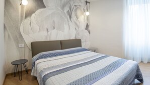A bedroom with a large bed, a wall mural, two pendant lights, a small round table, and a window with curtains.