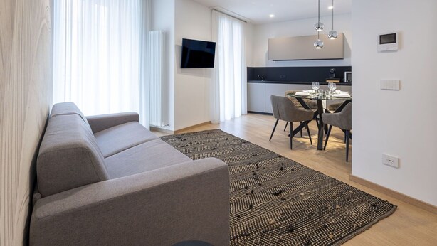 A modern living room with a sofa, a dining area with a table and chairs, a kitchen with a TV, and a large window with curtains.