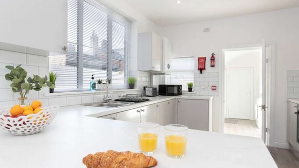 Une cuisine moderne avec des meubles blancs, un évier à deux bacs et un coin repas où l’on peut prendre le petit-déjeuner avec des croissants et du jus d’orange.