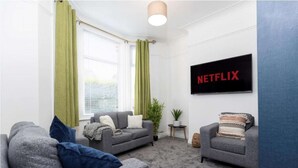 Un salon avec deux canapés gris, une table basse, un téléviseur fixé au mur et une fenêtre ornée de rideaux verts.