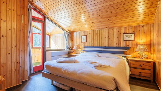 Un dormitorio tipo cabaña de madera con una cama grande, una mesita de noche con lámpara y una ventana con cortinas.