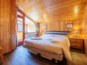 A wooden cabin bedroom with a large bed, a nightstand with a lamp, and a window with curtains.
