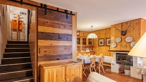 Interior acogedor de una cabaña con una escalera de madera, una zona de comedor con mesa y sillas, una chimenea y una cocina con estufa y armarios.