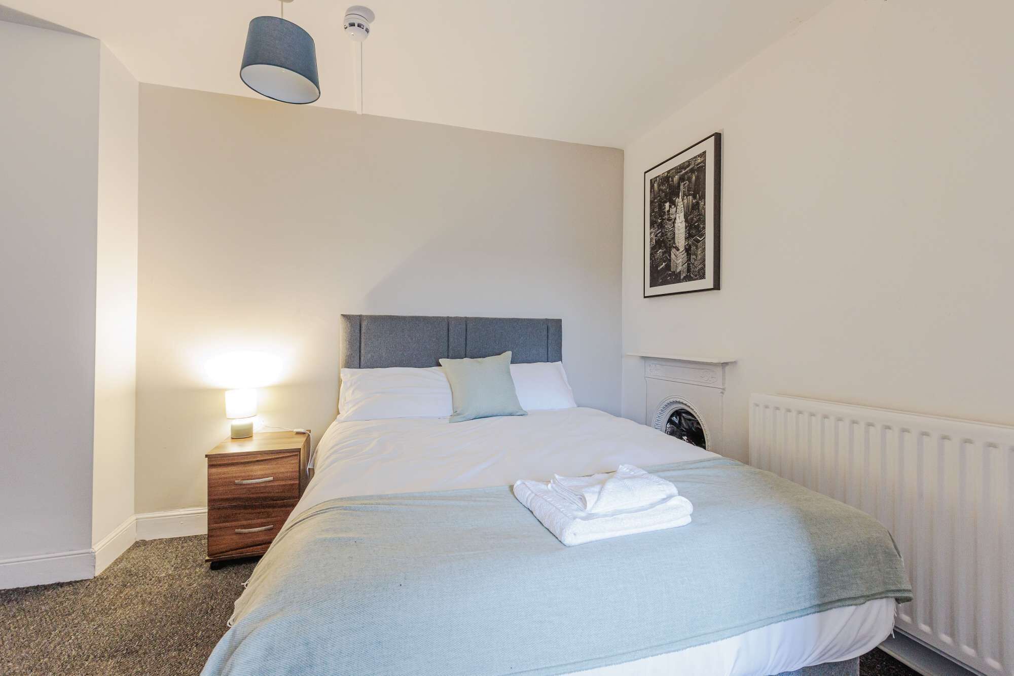 A neatly made bed with a grey headboard, a bedside table with a lamp, and a radiator on the wall.