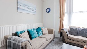 Ein Wohnzimmer mit einem beigefarbenen Sofa, einem gemusterten Sessel, einer blauen Uhr und einem Fenster mit Vorhängen.