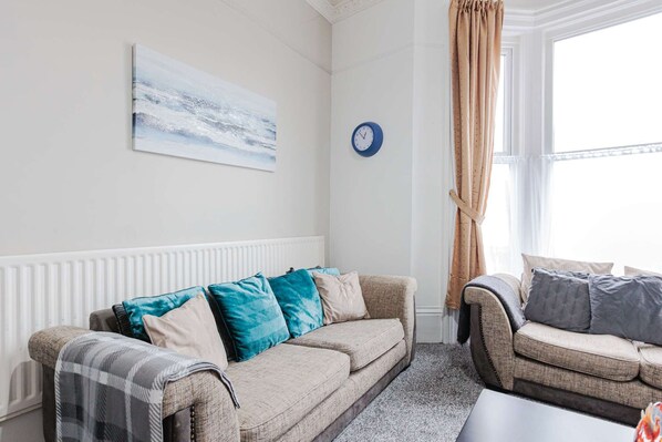 A living room with a beige sofa, a patterned armchair, a blue clock, and a window with curtains. - 5 Bedroom House in Hartlepool (Hartlepool)