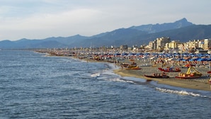 Une plage côtière bordée de nombreux parasols et de bateaux, un ciel dégagé et des montagnes en arrière-plan.