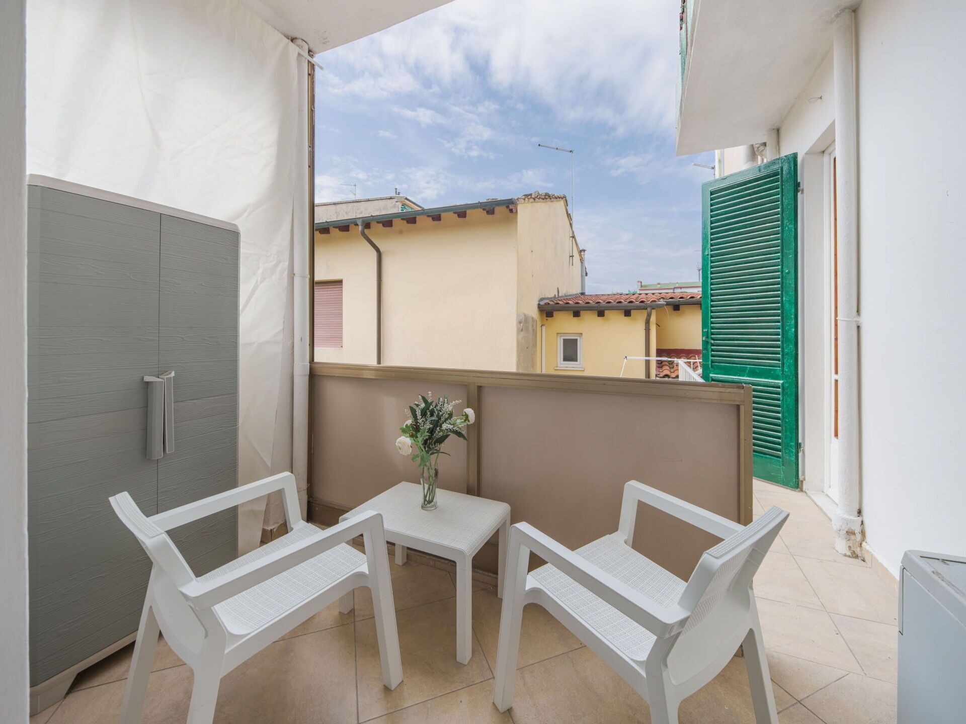 Ein Balkon mit einem weißen Tisch und zwei weißen Stühlen, einer kleinen Vase mit Blumen und Blick auf umliegende Gebäude.