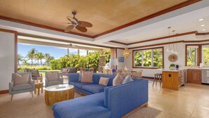 A spacious living room with a large blue sectional sofa, a wooden ceiling fan, and a view of palm trees through large windows. - Walk to MK Beach! 🏝️ Private Pool 🏊‍♂️ Jacuzzi ♨️ Mauna Kea Resort (Waimea)