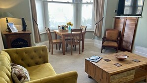 Ein Wohnzimmer mit gelber Couch, Esstisch aus Holz, Stühlen, einem Couchtisch aus Holz und einem Fernseher auf einem Schrank.
