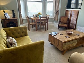 A living room with a yellow sofa, wooden dining table, and chairs, a wooden coffee table, and a television on a stand.
