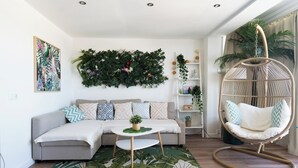 A modern living room with a grey sofa, a round wooden coffee table, a hanging chair, a large leafy wall hanging, and a bookshelf with plants. - Castelldefels - Apartment - Sleeps 4 - Beach Side (Castelldefels)
