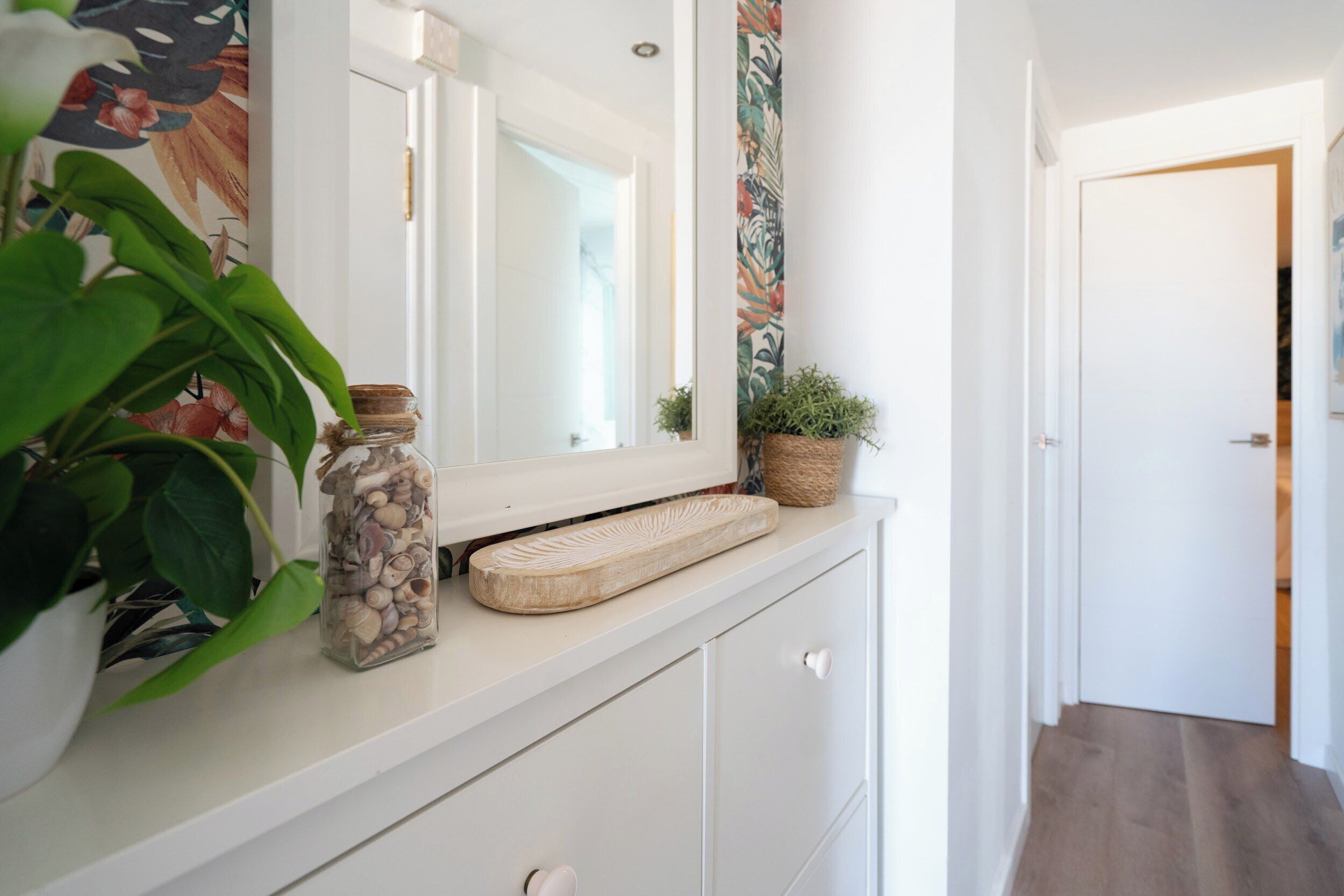 Een gang met een witte dressoir, een spiegel, een plant en een decoratieve mand.