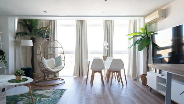 A modern living room with a round wooden table, white chairs, a hanging chair, a flat-screen TV, and large windows with curtains. - Castelldefels - Apartment - Sleeps 4 - Beach Side (Castelldefels)