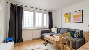 Ein modernes Wohnzimmer mit einem grauen Sofa, einem Holztisch und einem blauen Sessel. Ein Fenster mit schwarzen Vorhängen, eine Heizung und zwei gerahmte Bilder sind an der Wand zu sehen.
