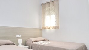 Habitación de hotel con dos camas individuales, cabecera de madera, mesita de noche con lámpara y ventana con cortinas traslúcidas.