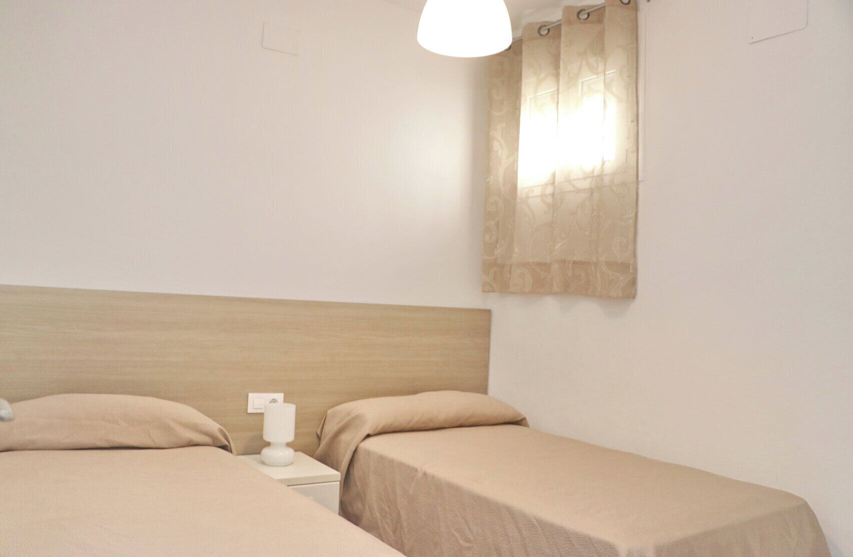 A simple room with two single beds, a wooden headboard, a nightstand with a lamp, and a window with curtains.