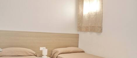 A simple room with two single beds, a wooden headboard, a nightstand with a lamp, and a window with curtains.