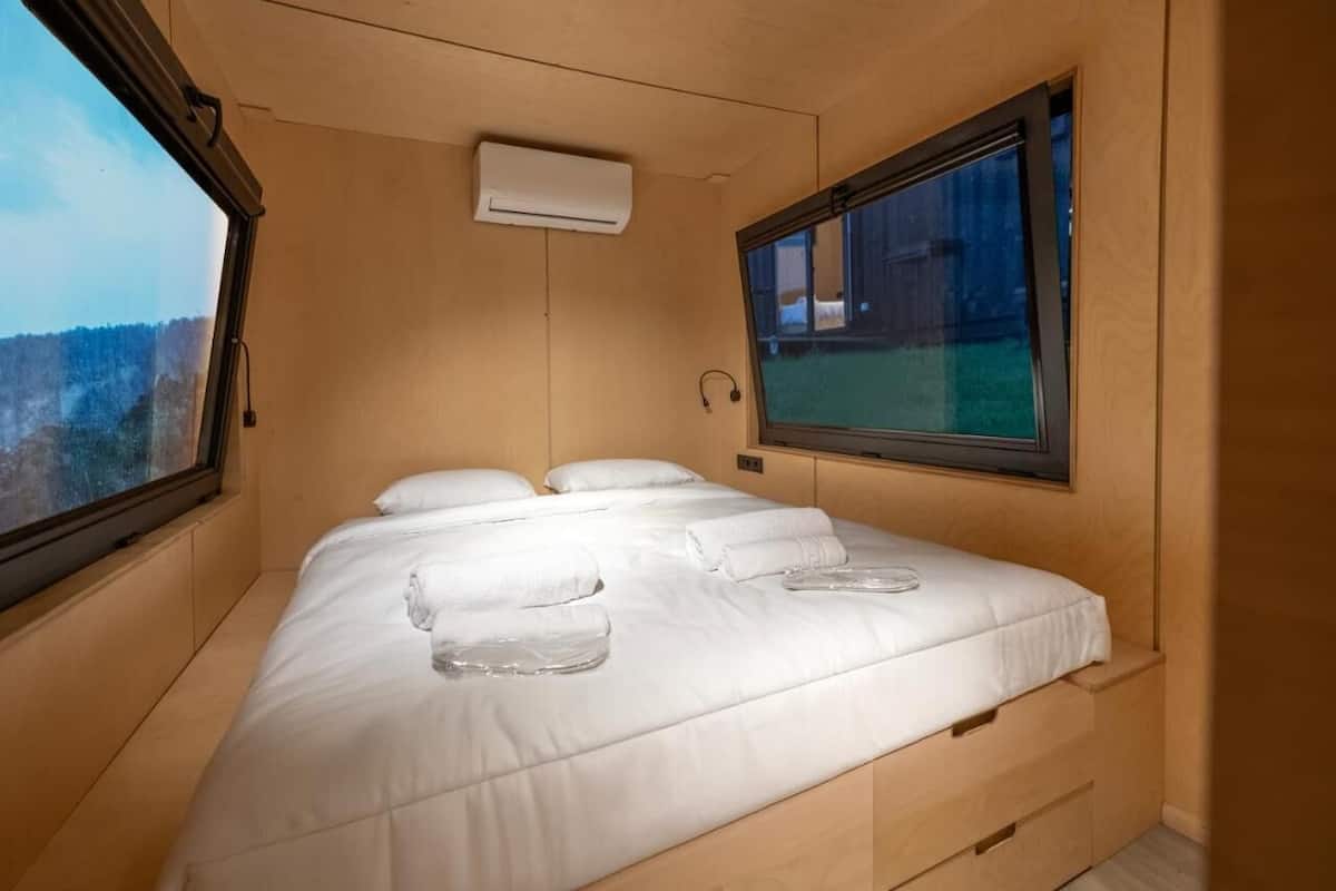 A single bed with white linens and towels, a window with a view of a building, and an air conditioning unit on the wall.