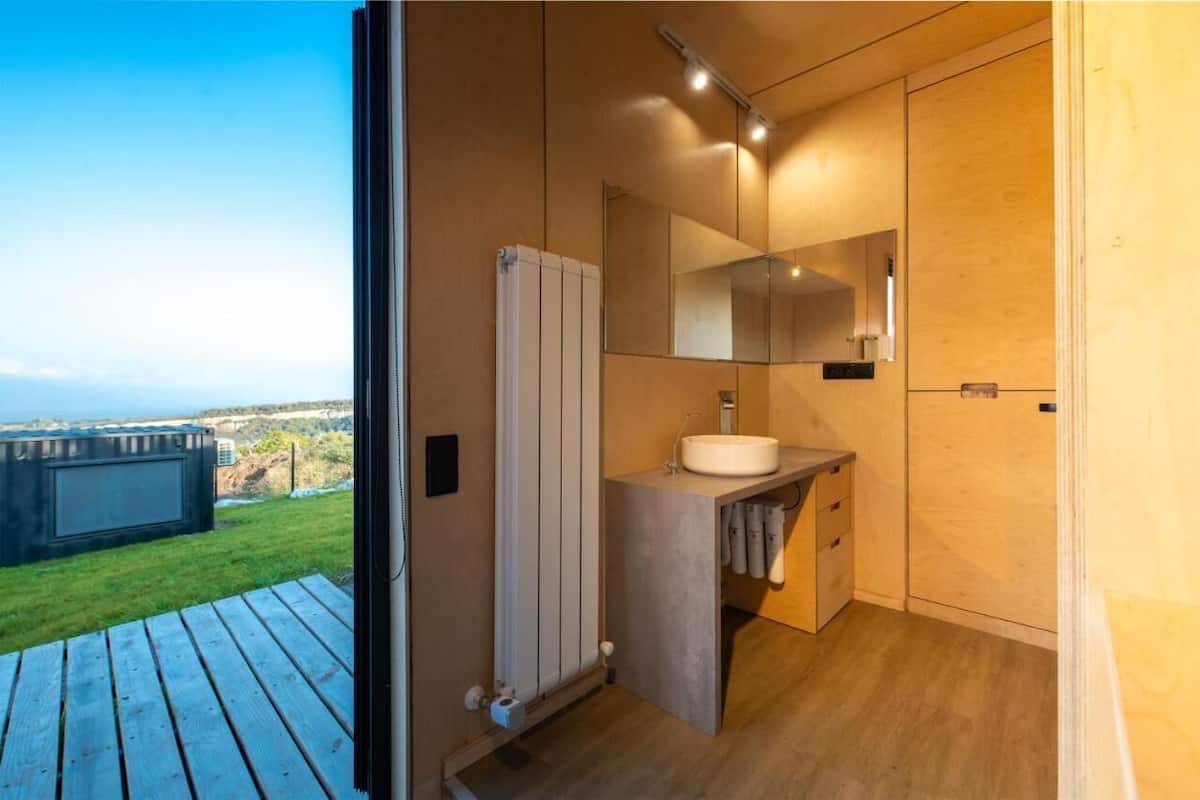 A modern bathroom with a large mirror, a sink, and a toilet. There is a wooden floor and a view of the outdoors.
