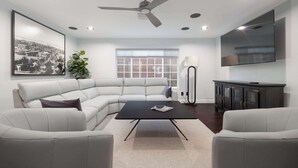 A modern living room with a sectional sofa, a black coffee table, a ceiling fan, recessed lighting, a large window, a floor lamp, a cabinet, and a flat-screen TV. - Modern Luxury | Hot Tub | 4 Blocks to Mission Bay! (San Diego)