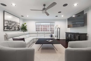 A modern living room with a sectional sofa, a black coffee table, a ceiling fan, recessed lighting, a large window, a floor lamp, a cabinet, and a flat-screen TV.
