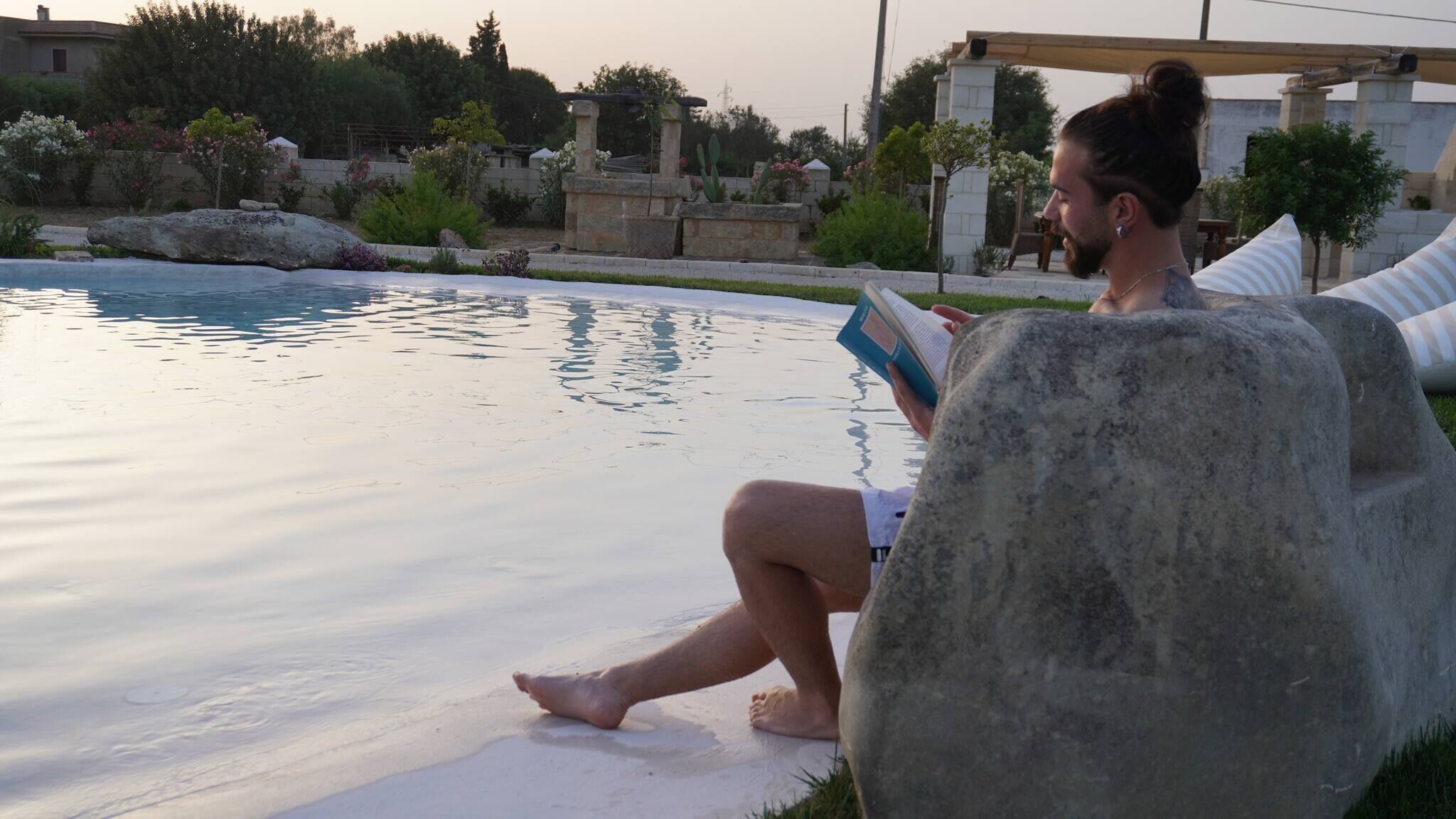 Una persona sentada junto a una piscina leyendo un libro, rodeada de un muro de piedra y follaje.
