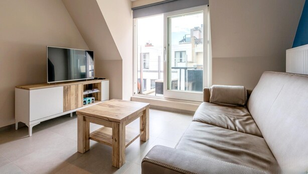 Een modern appartement met een loft-plafond, een flatscreen televisie op een houten kast, een beige bank en een groot raam met uitzicht op de stad.