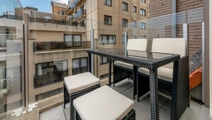 Een balkon met een glazen tafel en twee stoelen, met uitzicht op een gebouw met meerdere verdiepingen en balkons.