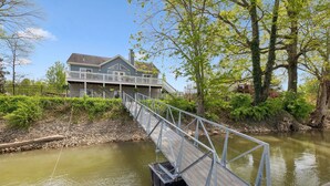 A house with a wooden deck extending over a river, surrounded by greenery and trees.