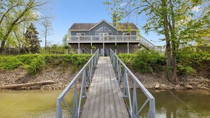 A wooden pier leading to a house with a balcony, surrounded by greenery and a river.