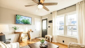 Ein Wohnzimmer mit Deckenventilator, einem an der Wand montierten Fernseher, einem Couchtisch aus Holz mit einer Pflanze und einem Fenster mit Vorhängen.