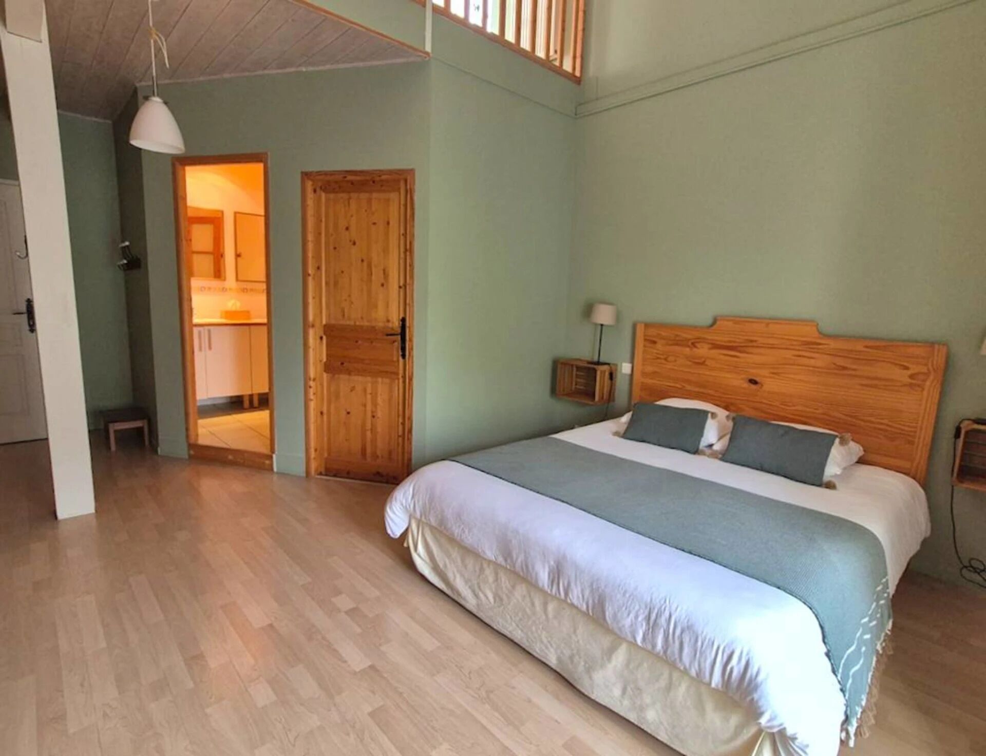 Un dormitorio con cabecera de madera, una cama grande con colcha blanca y azul, una puerta de madera y una mesita de noche con lámpara.