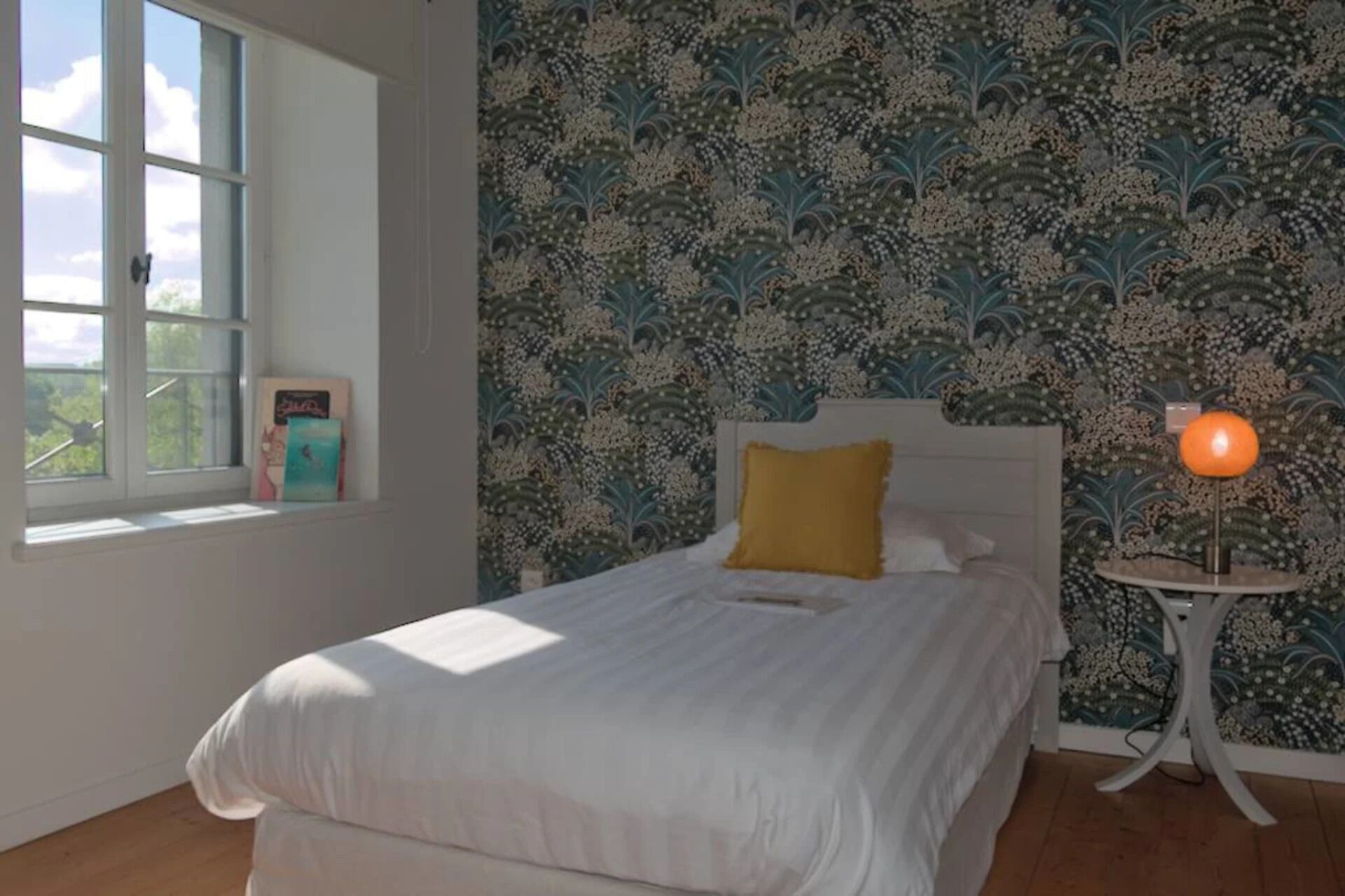 A bedroom with a bed, a nightstand, a lamp, and a window with artwork.