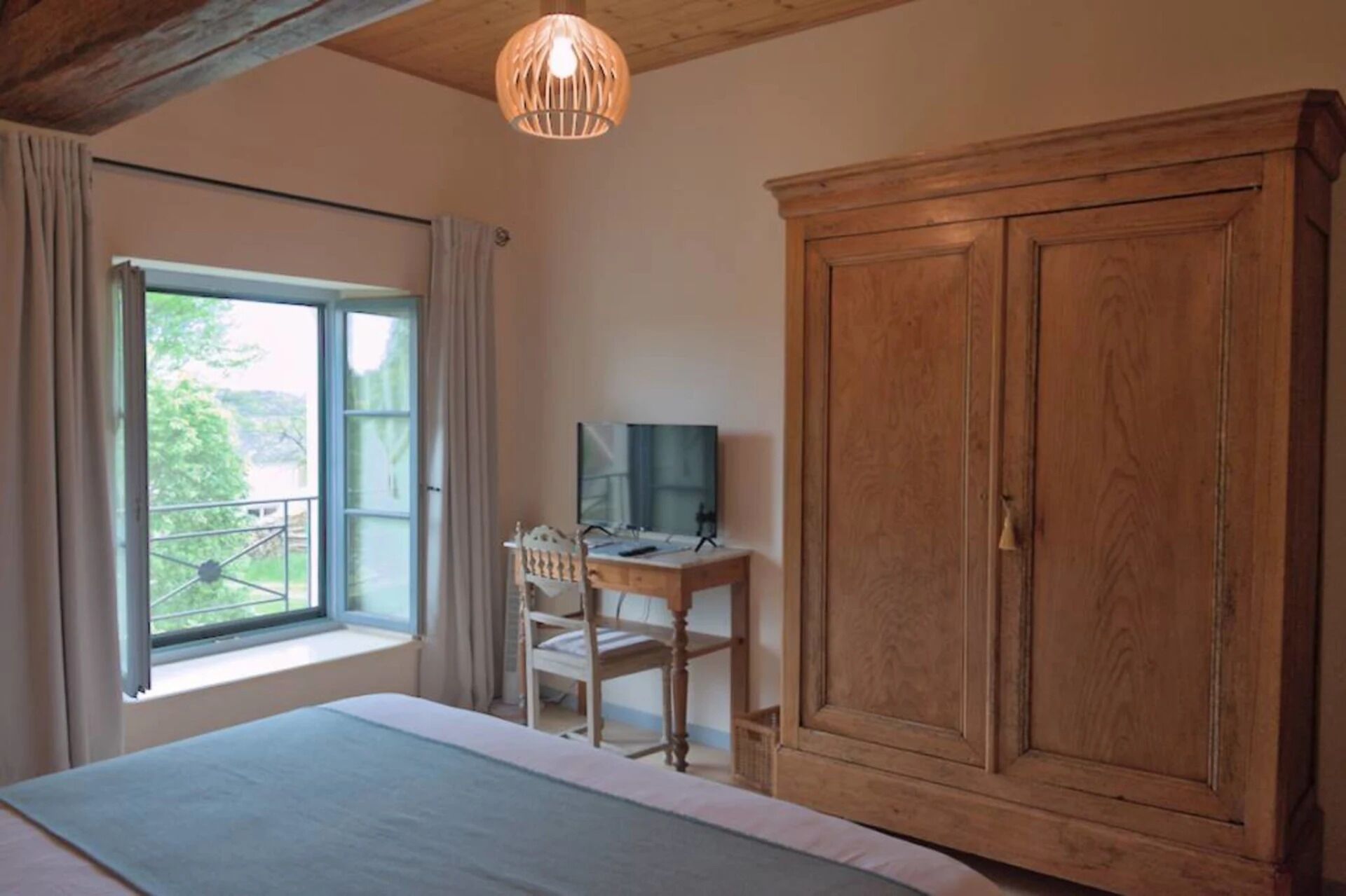 A bedroom with a large bed, a wooden wardrobe, a desk with a chair, and a window with curtains.