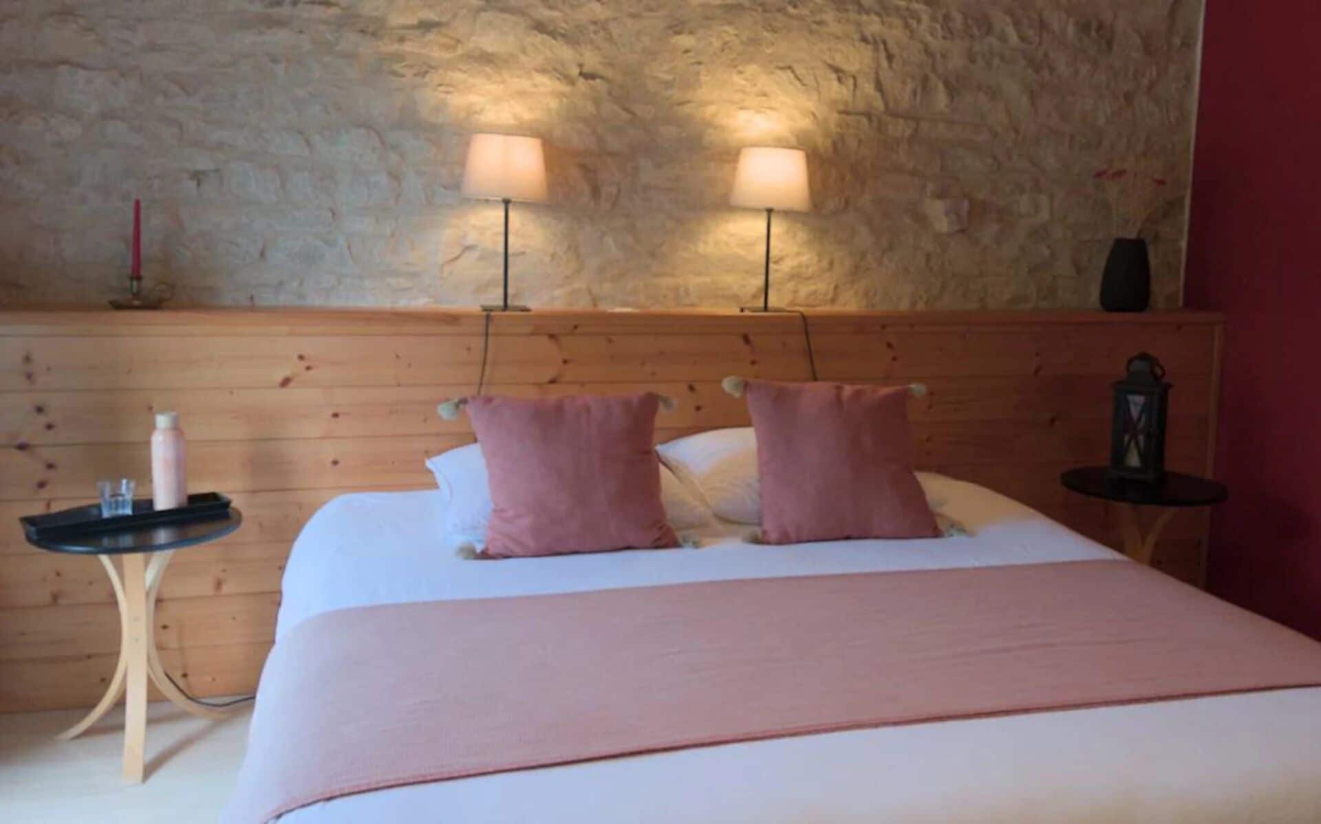 A bedroom with a wooden headboard, two bedside lamps, a bedside table with a bottle and glass, and a bed with pink pillows.