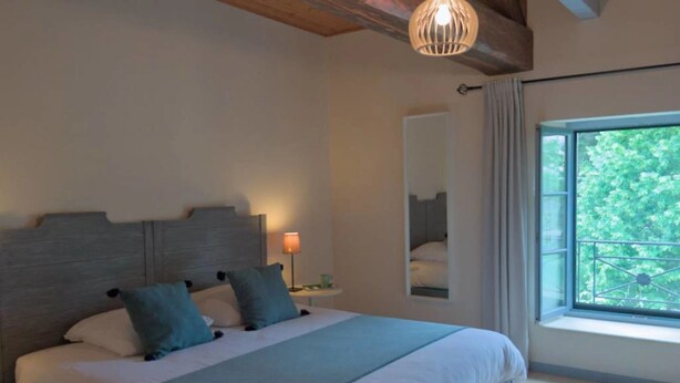 A bedroom with a large bed, a bedside table with a lamp, a window with a view of greenery, and a mirror on the wall. - Le Tilleul de Ray (Ray-sur-Saone)