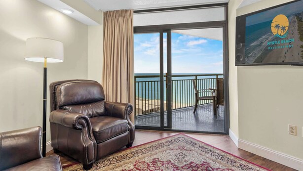 A room with a leather armchair, a floor lamp, a patterned rug, a sliding glass door leading to a balcony with a view of the sea, and a framed picture on the wall. - Sand Dunes Resort Unit 1406 - Ocean View! (Myrtle Beach)