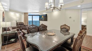 A dining area with a glass-top table, ornate chairs, a chandelier, and a view of the ocean through a large window. - Sand Dunes Resort Unit 1406 - Ocean View! (Myrtle Beach)