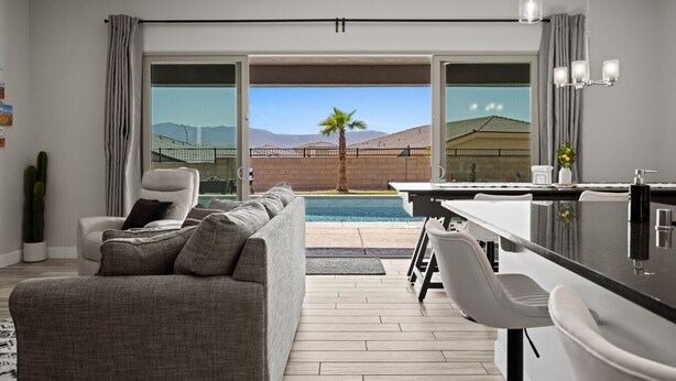 A modern living room with a grey sofa, a dining area with a bar, and a pool view.
