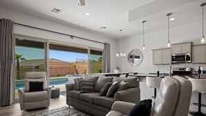 A modern living room with a gray sofa, a leather armchair, and a dining area with a kitchen in the background.